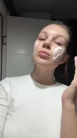 A fair-skinned young woman applies a white, creamy substance to her face in a bathroom setting. She is wearing a plain white T-shirt. The woman uses both hands to spread the substance across her face, including her forehead, cheeks, nose, and chin. The background shows a portion of a light-colored door and a darker area that may be a wall. The woman makes an exaggerated "O" shape with her mouth mid-application, suggesting she might be surprised or making a funny face. The scene is well-lit, and the camera is relatively close to the woman.
