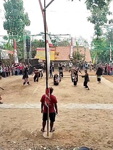 The video shows a traditional Indonesian performance in an outdoor, sandy arena. Several performers, dressed in traditional costumes and some with masks, are engaged in a dynamic, martial arts-like dance. One performer in the foreground, wearing a red shirt and shorts, stands facing away from the camera, observing the performance. In the background, an audience is gathered, watching the event. Flags, including the Indonesian flag, are visible, suggesting a festive or ceremonial occasion. Stage lights and speakers are set up, indicating a formal presentation. The environment appears to be rural, with trees and simple buildings in the distance. The overall atmosphere is one of cultural celebration and community gathering.