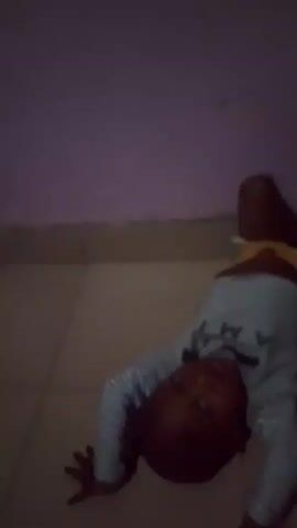 The video shows a young child lying on their back on a tiled floor. The child is wearing a light-colored t-shirt with black and white striped sleeves and yellow shorts. The child's head is tilted back, and their body is contorted as if they are attempting to do a handstand or bridge. The background appears to be a room with a purple wall and a tiled floor. The lighting is dim, suggesting it may be evening or nighttime. The child's facial expression is not clearly discernible due to the dim lighting and the angle of the shot. There is no discernible text in the video. The location is contextually identified as Yenegoa, Nigeria. The activity appears to be playful physical exertion or practice of a gymnastic movement.