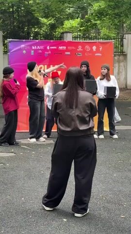 The video captures an outdoor event with a group of young people standing in front of a large banner. The banner is vibrantly colored, displaying a gradient from purple to orange and featuring various logos and text, some of which are in Russian. The visible Russian text reads "ДЕНЬ МОЛОДЁЖИ" (Youth Day) and "2023" indicating the event's nature and year. The backdrop suggests a public gathering or festival.

The foreground features a young person wearing a dark jacket and loose black pants, holding a microphone and facing away from the camera, seemingly addressing the group or performing. Behind them, five other individuals are positioned in front of the banner. Two are holding black square objects, one of which is a speaker or a similar electronic device. The group appears to be engaged in some sort of performance or presentation, as indicated by the microphone and the attentive stances of the individuals.

The setting is outdoors, with trees visible in the background and a textured ground surface, possibly asphalt or a similar outdoor flooring. The lighting suggests it is daytime, likely late morning or afternoon, with diffused light and shadows indicating overcast or shaded conditions. The atmosphere seems lively and youthful, with expressions ranging from focused attention to enjoyment. The presence of a microphone and the presentation setup points towards an organized event, possibly a talent show, speech, or promotional activity related to "Youth Day" in Dmitrov, Russia. The overall scene conveys a sense of community engagement and youthful energy.