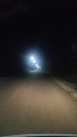 The video shows a view from a moving vehicle at night. The headlights of the vehicle illuminate a dirt road that appears to be in a rural area. In the distance, there are several bright lights, possibly streetlights or lights from a building. There is also a faint, illuminated cross visible amongst the distant lights. The sky is dark, indicating it is nighttime. There are no people or other distinct objects clearly visible besides the road and the distant lights. The atmosphere of the video is somewhat mysterious and desolate due to the darkness and the isolated nature of the setting. No text is visible in the video.