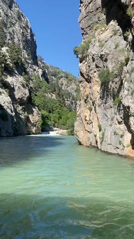 The video showcases a serene natural landscape, likely a canyon or gorge, with a vibrant turquoise river flowing between towering, rugged rock cliffs. The camera appears to be moving on the water, providing a dynamic view.

**Subjects:** The primary subjects are the steep, imposing rock cliffs that flank the river, along with the clear, flowing water. Sparse green vegetation, including trees and shrubs, clings to the rocky surfaces and is visible on the distant slopes.
**Activity:** The video captures a journey through the canyon, with the camera panning upwards to highlight the impressive scale and texture of the cliff face.
**Notable Details:** The time of day is bright, with a clear blue sky, suggesting sunny weather. The water has a distinct light green or turquoise hue, indicative of mineral content often found in such geological formations. The cliffs display various shades of grey, brown, and ochre, with visible cracks and stratification. The sparse greenery adds contrast to the rugged rock. The location is in Akseki, Turkey, known for its scenic natural beauty. No people or visible text are present in the video.