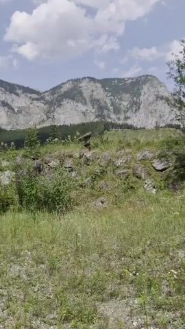 The video displays a wide shot of a mountainous landscape on a partly cloudy day. The foreground consists of gravel and green grass. In the mid-ground, there is a rocky incline with various shrubs and small trees, including some with purple flowers. A wooden fence structure is visible on the left side of the mid-ground, and beyond it, a building or structure can be seen. The background is dominated by large, steep mountains with visible rock faces and patches of trees. The overall impression is of a natural, somewhat rugged environment.