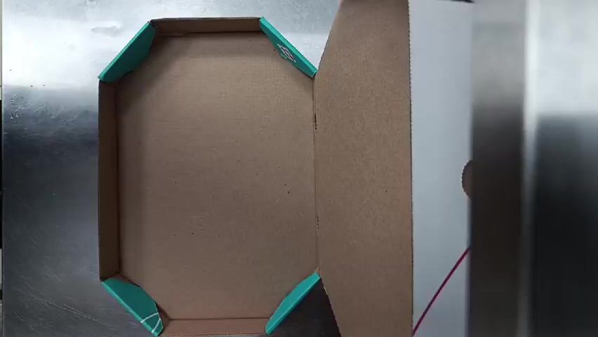 This is a top-down, slightly angled video of a rectangular pizza being placed into a pizza box. The pizza box is partially open, with the lid propped up. The inside of the box is made of corrugated cardboard, and there are four teal-colored triangular corner supports inside the base of the box. The pizza is on a sheet of brown parchment paper. It has a white crust, and toppings that appear to include bacon, green onions, and a yellow cheese. The pizza is placed into the box from the left side of the frame, and the parchment paper is adjusted slightly to fit. The background appears to be a metal surface.