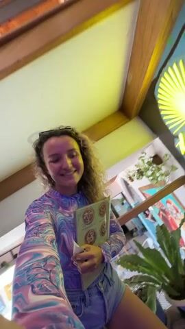 A young woman with curly brown hair, wearing a colorful tie-dye long-sleeved shirt and denim shorts, smiles as she holds a menu. She is standing in what appears to be a restaurant or cafe with wooden paneling on the ceiling and walls. To her right, a mural of cherry blossoms adorns a blue wall. A neon sign that reads "Blue Rays" is also visible. The camera, presumably held by the woman herself, is pointed at her face, capturing a selfie-style shot. The background shows glimpses of tables and chairs, suggesting an indoor dining area.
