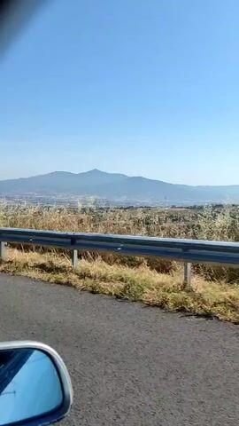 The video shows a first-person perspective from inside a car driving on a paved road. Initially, the view is from the passenger side window, capturing a blue side mirror in the bottom left, a metal guardrail, dry vegetation, and distant mountains under a clear blue sky. The camera then quickly pans forward, briefly showing the car's interior trim, before settling on a view through the front windshield. The car is on a multi-lane highway with a solid white line on the right and a broken white line in the middle. A metal guardrail separates the road from dry grassy land and scattered green bushes. Mountains are visible in the distance beneath a clear, bright blue sky. The car appears to be moving at a steady pace.