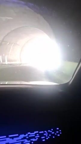 The video begins inside a tunnel, facing out toward the exit, with bright light obscuring the view. As the car emerges, a road stretches out toward massive, snow-capped mountains under a partly cloudy sky. The car is likely driving on a paved road toward the mountains.