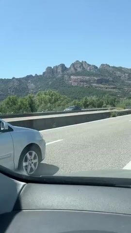 The video begins with a close-up shot of the rear of a silver Volkswagen Polo driving on a highway. The car's license plate reads FT 517 NK. The camera then pans out to show the car from a slightly higher angle, continuing to drive on the highway. In the background, a mountainous landscape with green vegetation is visible under a clear blue sky. The highway is paved with asphalt and has clear white lane markings. Other vehicles are visible in the distance on the highway, some in the same lane and others in adjacent lanes. The view is from inside another vehicle, with the dashboard and windshield frame visible in the foreground. The video continues to show the Volkswagen Polo moving further down the highway, with the mountainous scenery remaining consistent. The sun is shining brightly, casting shadows.