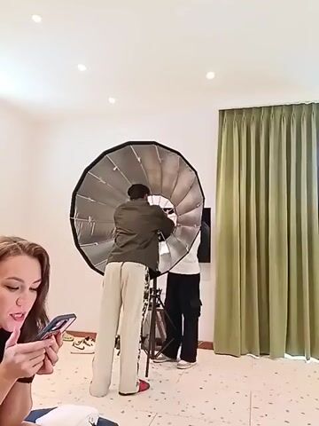 In a room with white walls, speckled flooring, and light green curtains, a woman with long brown hair is sitting to the left, looking at her phone. She is wearing a black top. Behind her, two men are setting up a large, silver reflector on a tripod. One man is wearing a green shirt and light-colored pants with red shoes, while the other is wearing a white shirt and dark pants. The room is well-lit with several ceiling lights. A TV can be seen in the background.