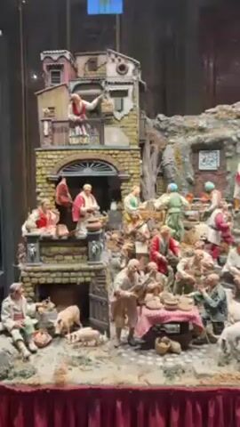 A detailed, multi-tiered diorama depicts a bustling scene of human activity, featuring numerous figurines arranged across various levels. The bottom tier includes figures sitting, standing, and interacting around a table laden with food, along with a pig and piglets. Above this, more figures are positioned on a structure resembling a building, engaging in various activities. Higher up, the diorama transitions into a rocky, mountainous landscape populated by more figures, some appearing to climb or gesture. Angels are positioned atop a small building, adding a religious element to the scene. The entire diorama is displayed behind glass.