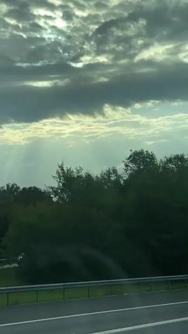 A video capturing a view from a moving vehicle, likely a car, shows a roadside scene. The foreground is dominated by a highway with a metal guardrail. Beyond the guardrail, a dense line of green trees stretches across the frame. The sky above is filled with dramatic clouds, with streaks of sunlight piercing through, suggesting either sunrise or sunset. A blue sign with the number 83.0 is visible near the guardrail. The overall impression is a transient, scenic view from a journey.