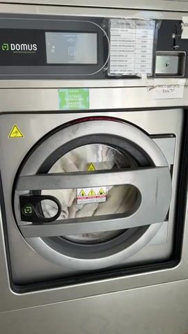 The video shows a close-up view of a commercial washing machine. The washing machine is made of stainless steel and has a large circular door in the center. Inside the drum, there are clothes visible. There are several labels and signs on the machine. One label, in green, reads "ULTIMA TECNOLOGIA CON UN CICLO DE DESINFECCION" which translates to "ULTIMATE TECHNOLOGY WITH A DISINFECTION CYCLE". Above the door, there is a control panel with a digital display and buttons. To the right of the control panel, there is a white paper with a list of programs and prices, seemingly for laundry services. The machine appears to be in a laundry facility or a similar commercial setting.