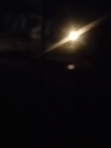 The video is a low-light recording of a dark outdoor scene. In the foreground, there are flashes of bright blue and red lights. In the background, there are blurred shapes and white lights, possibly headlights. The video appears to be shaky, suggesting it was recorded handheld while in motion. The overall impression is of a scene involving emergency vehicles or some sort of night activity with flashing lights.