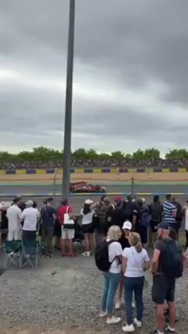 Here's a concise description of the video:

The video shows a crowd of people watching a race from behind a barrier at a racetrack. The sky is overcast. The race cars are moving quickly on the track. The crowd is casually dressed in light-colored clothing. In the background, more spectators are seated in stadium seating.