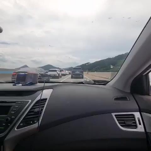 The video shows the interior of a car, looking out through the windshield. The car is in traffic, with several other vehicles visible ahead. The sky is overcast. The dashboard of the car is visible in the foreground. There is a phone mounted on the dashboard. The car appears to be on a highway.