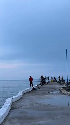 A group of people are fishing on a concrete pier. The pier is wet and has puddles of water.  There is a low, white wall lining the edge of the pier. The water is calm and stretches out to the horizon.  The sky is overcast and grey-blue. Most people are standing, with fishing poles in hand.  One person in a red jacket is standing on the wall, looking out at the water.  Fishing equipment, including buckets, is visible.