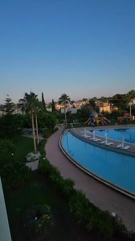 The video captures a panoramic view of a resort with multiple swimming pools and palm trees under a clear blue sky. The camera pans from left to right, revealing several connected pools, some with lounge chairs. In the background, there are buildings and what appears to be a water slide. The pools are filled with calm, blue water, reflecting the sky and surrounding structures. The scene is quiet, with no visible people in the immediate vicinity of the pools.