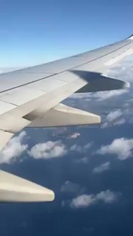 From the interior of a Ryanair passenger airplane, one wing is prominently displayed in the upper-left area of the frame. Looking down from the airplane, the ocean is visible, filled with puffy white clouds. Small islands, dark in color, can be seen in the distance. The sky is clear and blue.