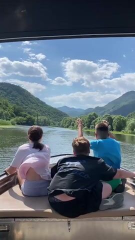 This media file captures a scenic boat ride on a river, likely in Riječani, Montenegro. Three individuals, two boys and one girl, are seated in the back of a boat, facing away from the camera and towards the stunning landscape. They appear to be enjoying a leisurely excursion.

The setting is a wide river, flanked by lush, green, tree-covered mountains that rise dramatically on either side. The sky above is a vibrant blue, dotted with fluffy white clouds, suggesting fair weather and likely daytime. The water is calm, reflecting the blue sky and surrounding greenery.

The individuals are casually dressed, indicating a relaxed atmosphere. The girl on the left has her hair in a ponytail and wears a light-colored shirt and shorts. The boy in the middle wears a black t-shirt and shorts, while the boy on the right sports a bright blue t-shirt and green shorts. One of the boys in the middle is actively gesturing with his hand, perhaps pointing out something of interest in the scenery or expressing excitement. The overall mood seems joyful and adventurous, with the children immersed in the beauty of their surroundings.

No discernible text is visible in the media file. The focus is on the natural beauty and the shared experience of the boat trip.