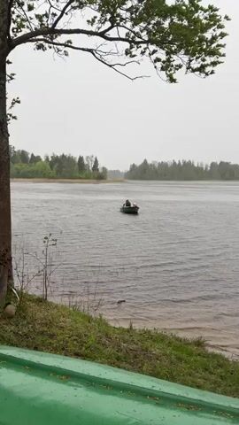 A video depicts two people in a small green rowboat on a body of water, likely a lake. One individual, appearing to be an adult, is wearing a dark jacket with the hood up and is sitting at the back of the boat. The second person, possibly a child, is in the front and wearing a bright orange life vest and a light-colored hat. The boat is equipped with a small electric motor at the back and oars. It is raining and the water is choppy. The shoreline is visible with a grass embankment and trees in the background. The sky is overcast, creating a grey, damp atmosphere.