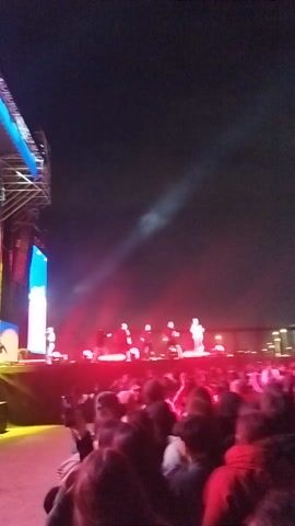 The video captures a lively nighttime event in Maipú, Chile, centered around a large outdoor stage. Bright, colorful stage lights, predominantly red, blue, and yellow, illuminate the scene. Two large screens are visible on the stage. The screen on the left displays the text "FIESTAS PATRIAS en Maipu" (Patriotic Holidays in Maipú) in white capital letters on a red and blue background with snowflake-like graphics. Below the text, an image of a smiling person, possibly a performer or host, is shown. The audience is visible on the right side of the frame, facing the stage and enjoying the event. The atmosphere appears festive and energetic, characteristic of a public celebration or concert.