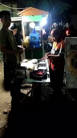 This video shows an outdoor scene at night, lit by a bright lamp. A food cart with a water dispenser is visible. Two women and a young man are interacting. One woman, wearing a headscarf, is handing something to another woman who appears to be making a transaction or preparing food. The young man stands to the side, observing. There are food items on display on a table in front of the cart. A sign with the word "MOGA" is visible on the right side of the frame. The ground appears to be dirt or gravel.