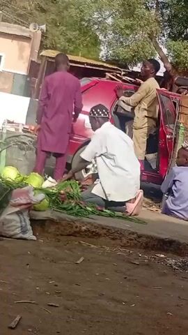 The video shows a street scene in what appears to be a market area. Several people are walking by. A red car with an open door is visible; it is surrounded by various items, including produce like cabbage and onions, as well as bags and boxes. Some people are near the car, while others are walking past it. One person is kneeling on the ground next to the produce. The environment includes trees, buildings, and what seems to be a dirt or gravel road.