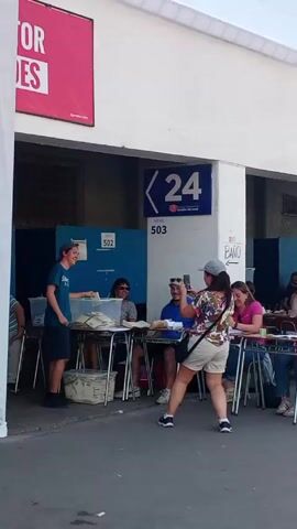 The video shows a polling place with people voting. A young man in a blue t-shirt and shorts places his ballot into a clear container on a table manned by several adults. A woman in a floral shirt and shorts takes pictures with her phone as she walks past the tables. There are signs indicating "Mesa 502" and "Mesa 503" above booths, and a larger sign with "24" on a blue background. A pink banner with Spanish writing hangs to the left. The setting appears to be outdoors, but covered, with white walls and concrete flooring.  People are seated at tables, some with paperwork in front of them, possibly poll workers.