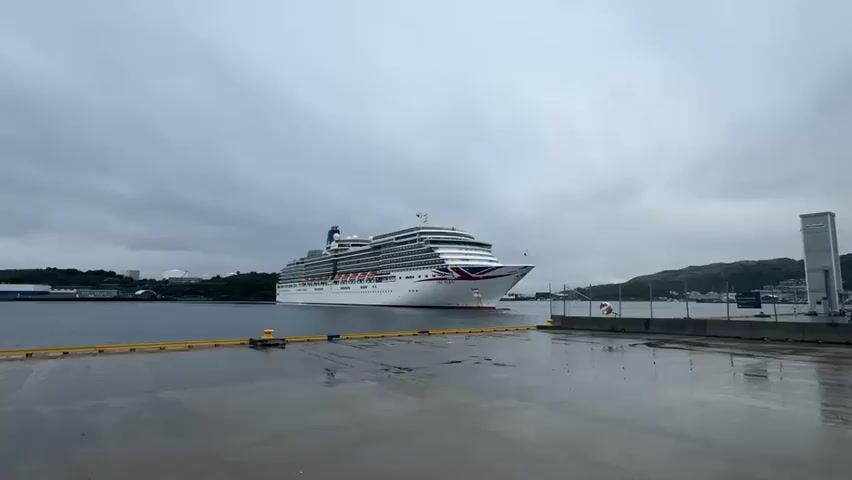 The video shows a large white cruise ship with a red and blue stripe on its hull sailing on the water. The ship is in the middle of the frame. In the foreground, there is a wet pier with yellow ropes and bollards. In the background, there are hills and buildings. The sky is cloudy and overcast.