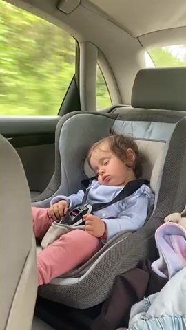 A close-up shot displays a toddler sleeping peacefully in a gray car seat. The child has fair skin, brown hair, and is dressed in a light blue, long-sleeved shirt and pink pants. The car seat is equipped with a black safety harness that secures the child. The background shows the interior of a car with a window partially visible, suggesting the car is moving as the scenery outside is blurred. The child's face is turned slightly to the side, revealing chubby cheeks and slightly parted lips. A pacifier clip is visible near the child's chest. The overall setting is intimate, capturing a quiet moment during a car ride.