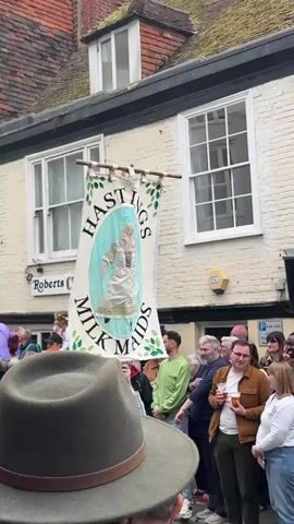 The video shows a procession of people in Hastings, UK, carrying a banner that reads "Hastings Milk Maids." The banner has a painting of a woman in traditional clothing. The crowd includes people of all ages, some in costume, and a few are holding drinks. The street is lined with old buildings, including a shop called "Roberts Curios." The weather appears overcast.