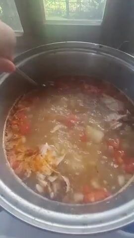 A close-up shot shows a pot of soup with tomatoes and meat simmering on a stove. Steam rises from the soup, and a spoon is used to stir it. The pot is placed on a white electric stove, and a window with a view of trees and a fence is visible in the background. The lighting suggests it is daytime.