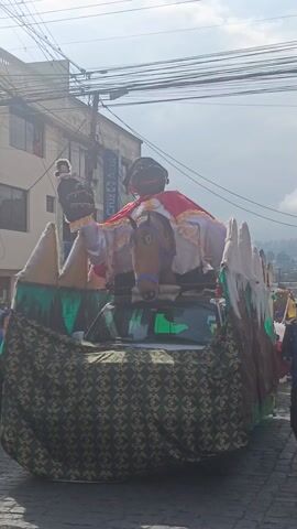 A slow-motion video shows a festively decorated vehicle moving down a cobblestone street. The vehicle is adorned with a large, puppet-like figure dressed in white and red, riding a hobby horse. The figure holds a smaller doll in its raised hand. The vehicle's base is covered in a dark, patterned cloth. In the background, onlookers are visible, and buildings line the street. Overhead, power lines crisscross the sky. The video is shot in daylight, but the sky is overcast. The overall atmosphere appears celebratory.