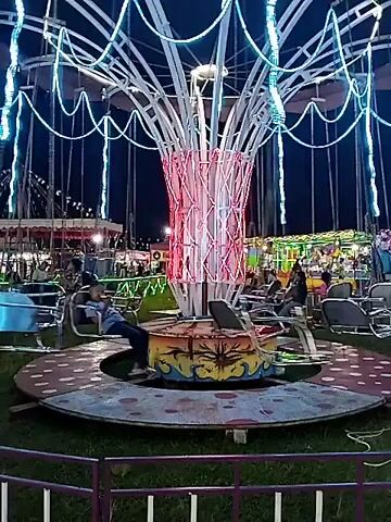 At night, an amusement park ride is in operation, featuring a central tower with a red illuminated latticework design. From the top of the tower, multiple cables with blue lights extend outwards and downwards, supporting small, airplane-shaped structures that rotate in a circle around the base of the tower. People are seated in the airplane structures as the ride is in motion. The ride is on grass and is surrounded by a low metal fence. In the background, other amusement park attractions can be seen with bright lights and activity. The sky is dark.