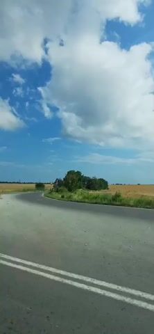 The video shows a car traveling on a road with agricultural fields on the right. A blue road sign with white lettering indicates directions to "Varna," "Yugol," "Kavarna," and "Balchik." The sky is blue with white clouds. A few trees are visible along the roadside. The road appears to be in a rural area.