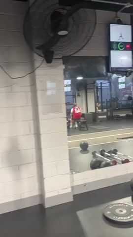 The video shows a gym. In the foreground, weights and medicine balls are visible on the floor. In the middle ground, a person in a red and white shirt is sitting on a bench, facing away from the camera, in front of a mirror. Behind the person, the gym equipment is reflected in the mirror, including weight racks and punching bags. Above the mirror, screens display exercise demonstrations. In the background, punching bags and other gym equipment are visible. The overall environment appears to be a modern fitness facility.