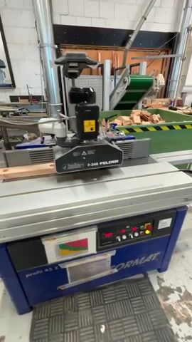 The video shows a woodworking machine in operation, specifically a Felder F-348. The machine has a blue base and a silver tabletop with a ruler marking. A wooden workpiece is being fed through the machine, and a cutting head is visible above it. The cutting head has a black motor and a warning label. A digital display on the machine shows the number 1000. In the background, a green conveyor belt is visible, carrying a pile of wooden blocks. The environment appears to be a workshop or factory, with brick walls and other machinery in the background. The video is a close-up shot of the machine in action, focusing on the woodworking process.