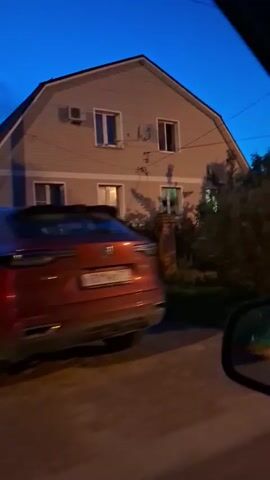 The video shows a view from a moving vehicle, likely a car, at dusk or night. The sky is a deep blue. In the foreground, a portion of the car's interior, including a side mirror and a red car, is visible. The main focus is on a residential street with several houses. The houses are light-colored, with visible windows, some of which are lit from within. There are trees and bushes in front of the houses, and some have brick fences or gates. A utility pole is also visible. The motion blur indicates the vehicle is in transit.