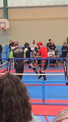 Two fighters in a boxing ring, one in red, the other in black, engage in a sparring session. Each wears a head protector, gloves, and shin guards. The red fighter is slightly heavier set than the black. They’re inside a boxing ring with blue mats and red borders, surrounded by blue ropes. Several spectators watch from beyond the ring, some seated at a table. A referee wearing a neon green vest observes the fight closely. The scene is indoors, possibly in a gym or event space with basketball equipment in the background, a large speaker is also present.