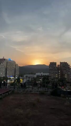 This vertical video captures a serene sunset over a city skyline in Les Amplaries, Spain. The sun, a bright orange orb, is setting behind a silhouetted mountain range, casting a warm glow across the sky. Wispy clouds streak across the horizon, tinged with hues of orange, pink, and gray.

The foreground is dominated by a dry, grassy field. Beyond this, the city unfolds with several multi-story buildings. A tall, white building on the left features the word "IBIZA" illuminated in blue letters near its top. To the right, a cluster of brown, apartment-like buildings stands. Between these structures, a street with palm trees lining its sides is visible, suggesting a Mediterranean or tropical climate. The presence of streetlights and building lights indicates the transition from day to evening. The overall mood is tranquil and picturesque, typical of a peaceful sunset in a coastal or resort area. The time of day is clearly sunset, and the weather appears clear with scattered clouds.