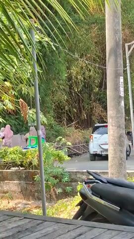 A white car is parked on a road next to a tall pole. A person wearing a dark shirt and pants is standing near the car. In the background, there is dense vegetation, including bamboo. In the foreground, there is a portion of a motorcycle. To the left, there are people wearing headscarves and some laundry is hanging.