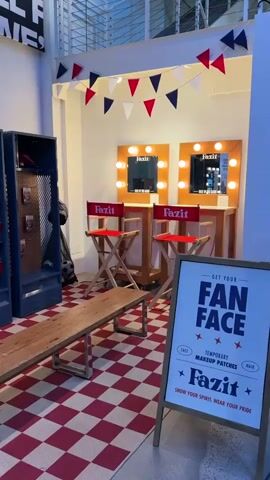 The video shows a retail display area. In the foreground, a red and white checkered floor is visible. To the left, there is a long wooden bench with metal legs. Behind the bench are three tall blue lockers with pegboards inside, displaying what appear to be temporary makeup patches or stickers with images of faces. A soccer ball is visible on top of the middle locker. Above the lockers, a sign reads "Fazit" with stars around it, illuminated by lights.

To the right of the bench, there are two vanity mirrors with lights around them, set on wooden stands. In front of these mirrors are two red director's chairs with "Fazit" printed on them.

In the far right of the scene, a sign is propped up that says "GET YOUR FAN FACE TEMPORARY MAKEUP PATCHES Fazit SHOW YOUR SPIRIT, WEAR YOUR PRIDE".

Further in the background, there are more display shelves with clothing items and possibly accessories. A staircase is visible in the reflection of the mirrors, and a person appears to be walking up it. The overall environment suggests a promotional space, possibly for a sports-themed event or brand. Decorative red, white, and blue pennant flags are strung across the top of the display.