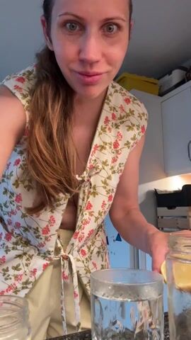 A woman with long brown hair is indoors, standing in front of a kitchen counter. She wears a white floral print top tied at the front and light yellow shorts. She squeezes a lemon half into a glass of water, stirs it with a spoon, and then empties the contents of a jar into the glass. The kitchen has white cabinets, and a yellow storage container is visible on top of one of the cabinets.