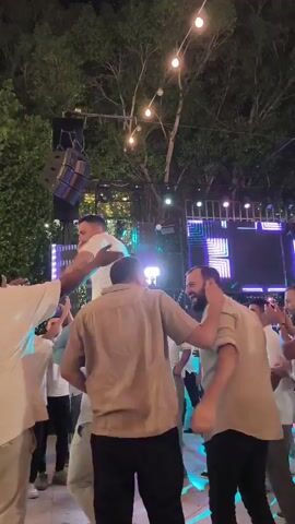 The video shows a group of men dancing outdoors at night. Several men are wearing white shirts and light-colored pants. One man is being carried on the shoulders of another man, and he is raising his arms in the air. There is a DJ booth with lights in the background, and string lights are hanging from trees overhead. The atmosphere appears festive, likely at a party or celebration.