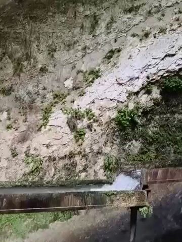The video shows a waterfall cascading down a rocky cliff face covered in lush green vegetation. At the bottom of the waterfall, a man-made structure, appearing to be a trough or channel made of metal and wood, directs the flowing water. The water is turbulent and frothy as it moves through the channel. The environment is natural, with the waterfall and vegetation dominating the scene, suggesting a remote or natural setting that has been partially engineered for water management.