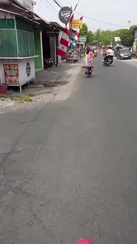 A person is riding a scooter down a street in a town. There are buildings on the left side of the street, and a few people are riding scooters and motorcycles on the right side. There are also a few flags and balloons on display. The weather is overcast.