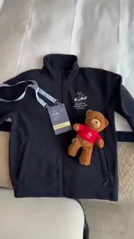 The video shows a jacket, badge, and teddy bear laying on a bed. The jacket is a dark color. The badge is hanging from a white lanyard, and has the word "DRIVER" printed on it. The teddy bear has brown fur and is wearing a small red shirt with a logo on it. The camera zooms in to show the logo on the shirt, then zooms back out.