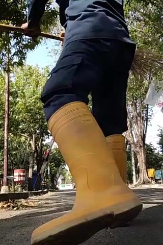 A person wearing a dark blue jacket, dark pants, and yellow boots is holding a broom upside down and walking away from the camera on a paved path. The person is also wearing a yellow hard hat and a dark fabric covering their face and head. Trees with green leaves line both sides of the path, and there are some red and white flags on poles in the background. The sky is visible and appears to be blue with some clouds. The ground in the foreground is a darker shade of asphalt, and there are some leaves and debris scattered on it. The video is shot from a low angle, looking up at the person and the broom.