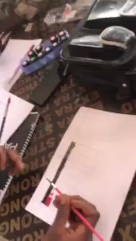 A video shows a person using a red marker to color a design on a piece of paper. The paper is laid on a table with a grid-like design on it. A ruler is being used to assist with the coloring. Another person is seen in the background, also drawing on a piece of paper. There are several items on the table including a pencil case, a bag, and other papers. The table has writing on it that includes the words "EXTRA STRONG".