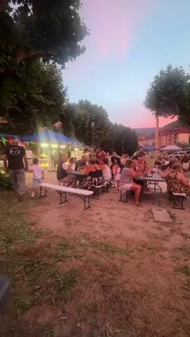 The video shows an outdoor gathering with people seated at tables under a twilight sky with pink and blue hues. There are trees and some structures that appear to be tents or booths in the background, possibly for a market or festival. The ground is covered in dirt and grass. Some individuals are standing and walking around, while others are engaged in conversation or eating. The overall atmosphere seems relaxed and social.